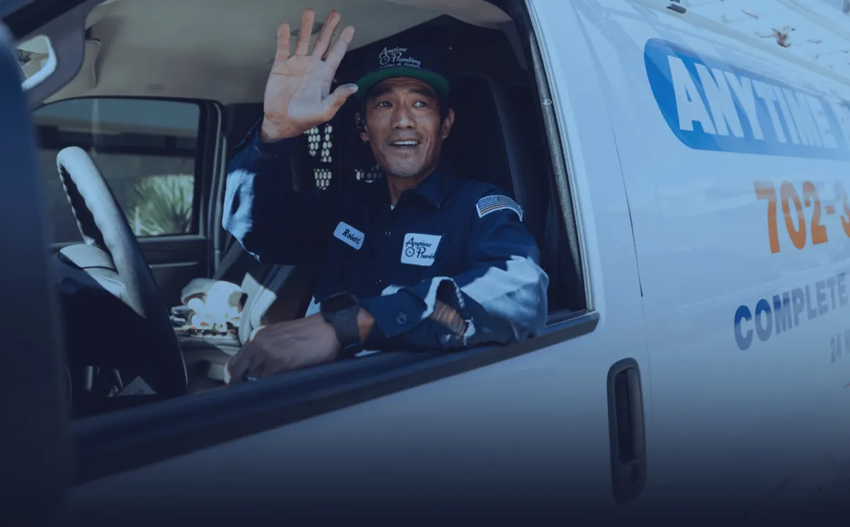 An Anytime Plumbing technician in a navy company uniform waving from the driver’s seat of a company van