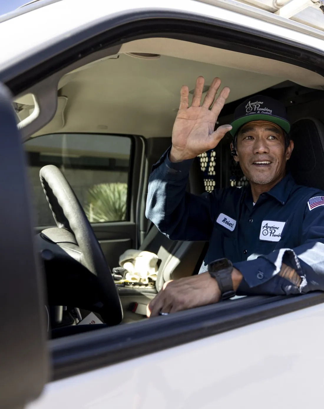 An Anytime Plumbing technician in a navy company uniform and cap waving from the driver’s seat of a lettered company van