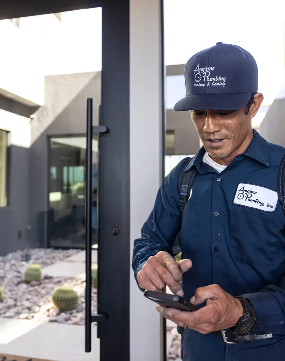 An Anytime technician in a navy uniform and cap at a front door, looking at his phone