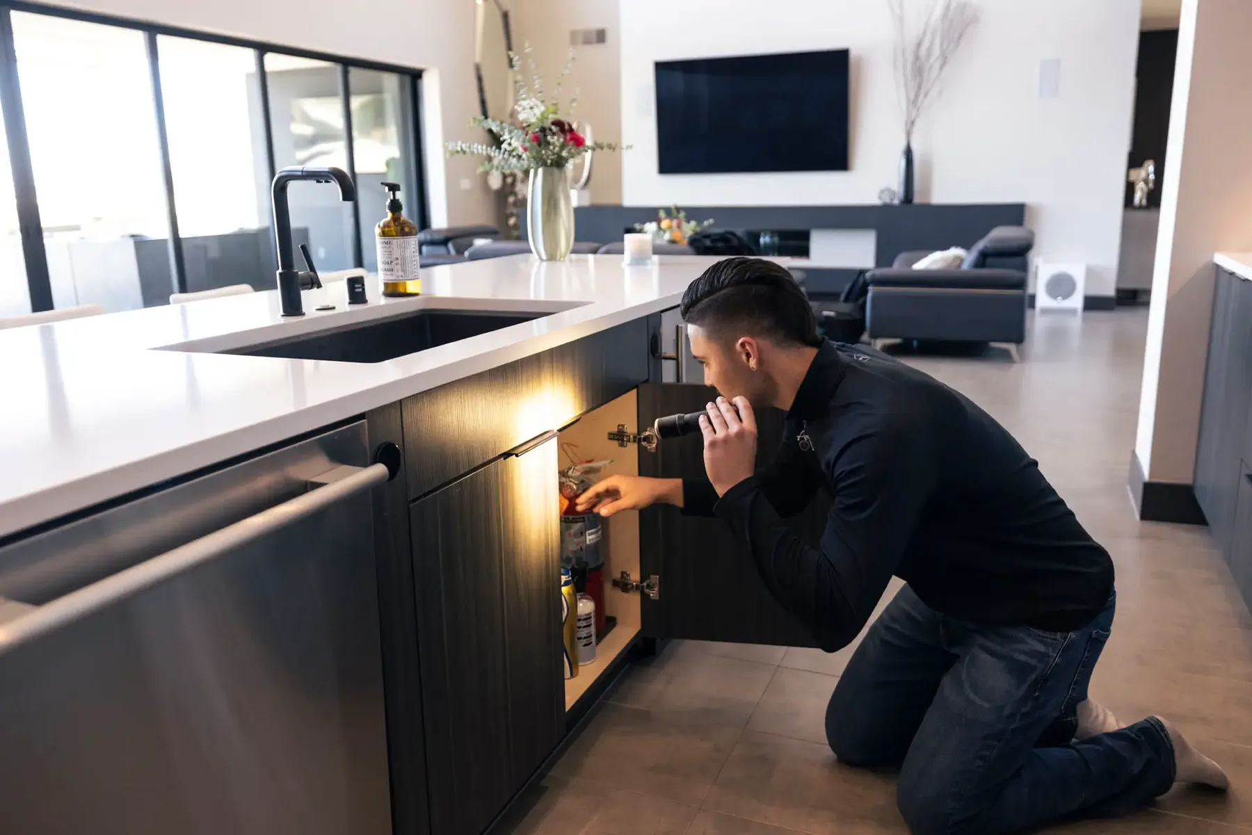An Anytime technician kneeling with a flashlight at an open under-sink cabinet in a modern kitchen