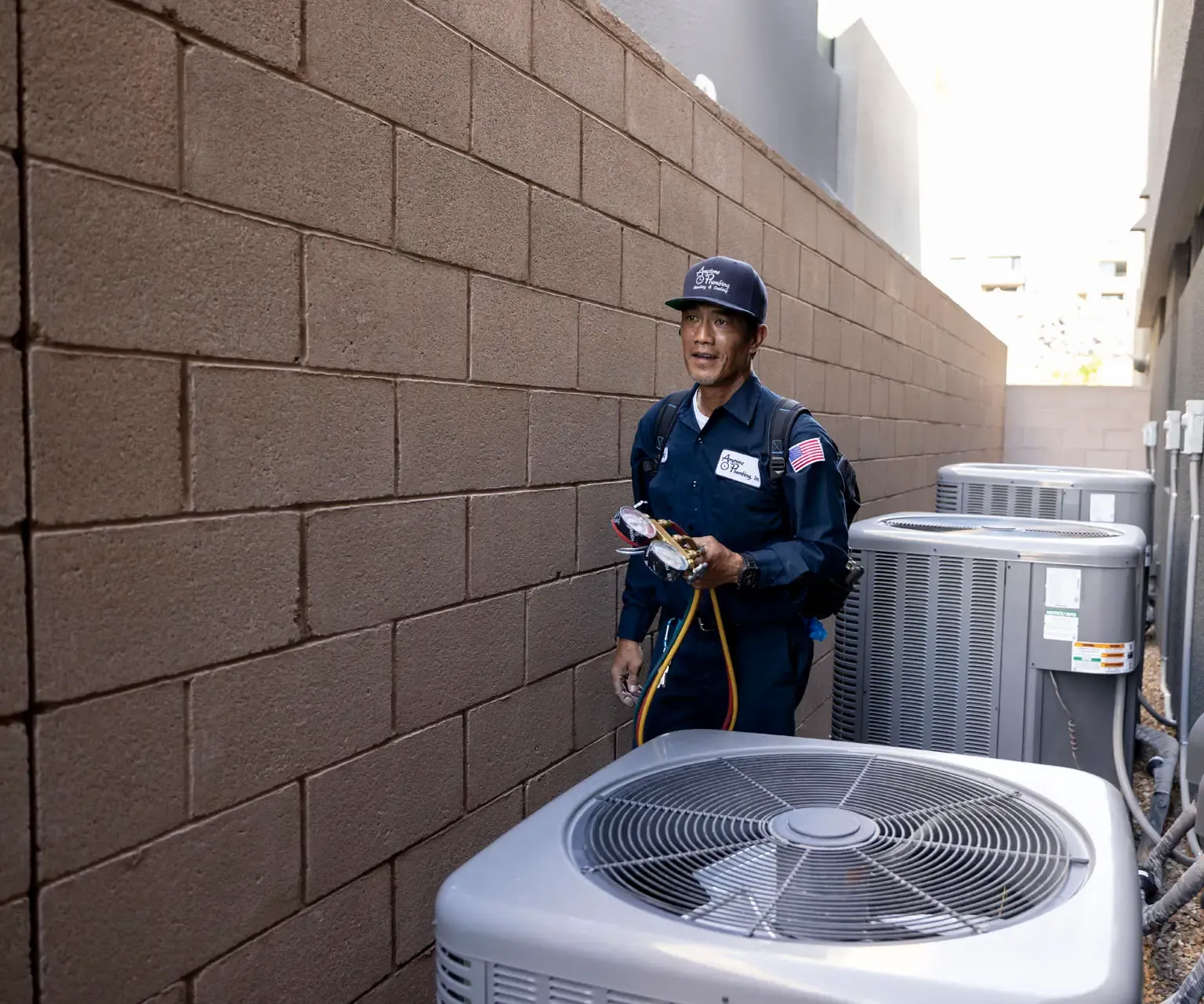 An Anytime technician in a navy uniform walking between block walls beside two outdoor AC condensers