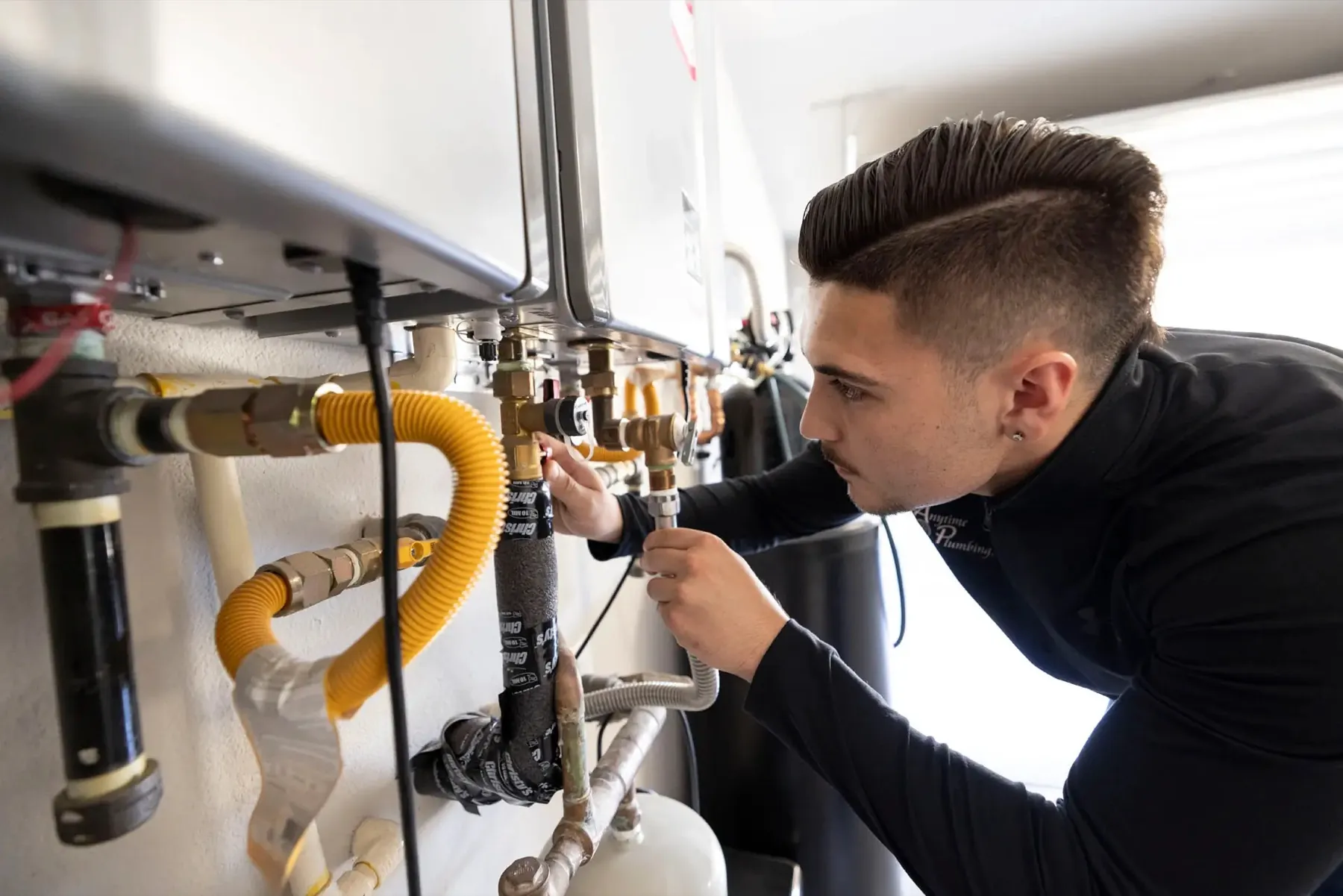 An Anytime Plumbing technician adjusting a valve beneath a pair of wall-mounted tankless water heaters