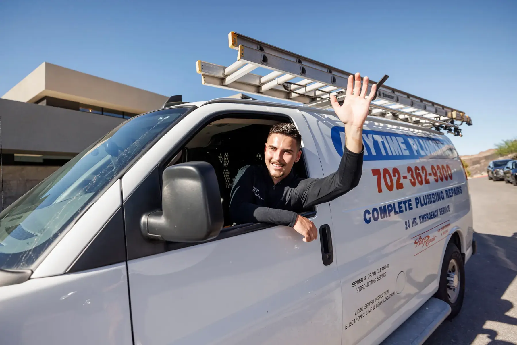 An Anytime technician waving from the window of a lettered company van with a ladder rack